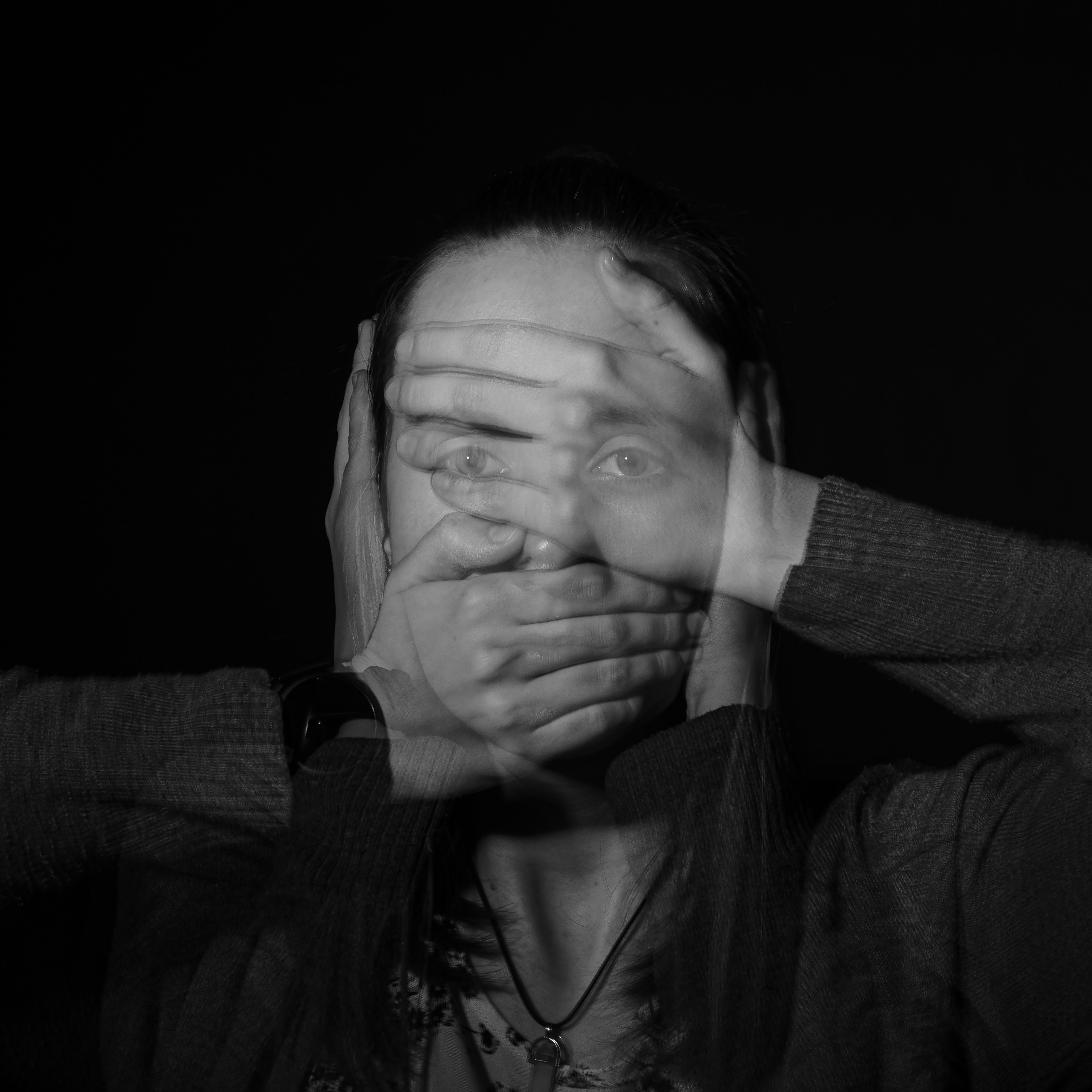 Person holding both hands to the sides of their head, wearing a necklace and sweater, set against a dark background with dramatic lighting creating a moody and introspective atmosphere