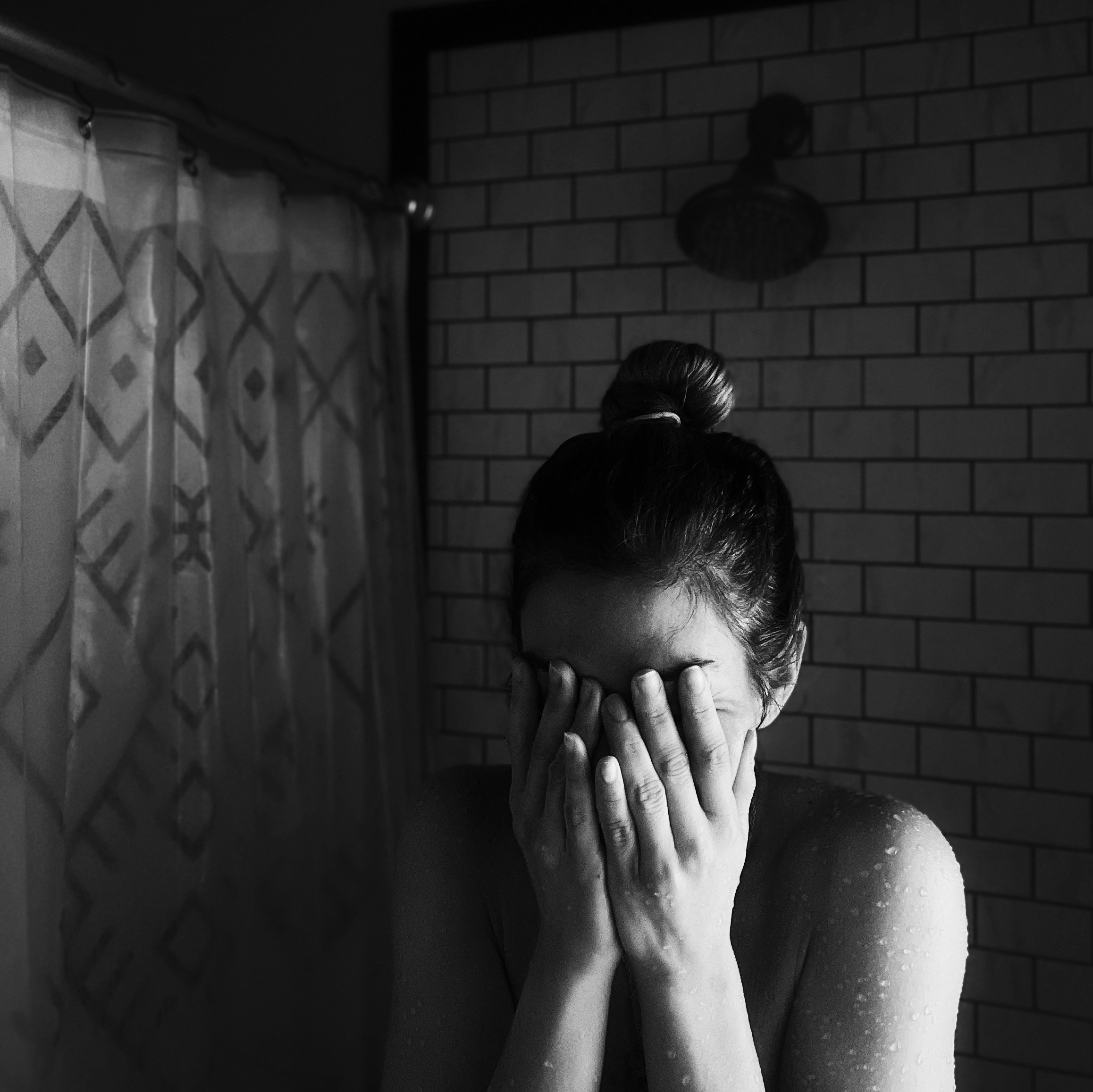 Person standing in a tiled shower with hair tied in a bun, covering their face with both hands, water droplets visible on their shoulders and arms, soft natural light creating a calm and introspective mood, patterned shower curtain and showerhead in the background