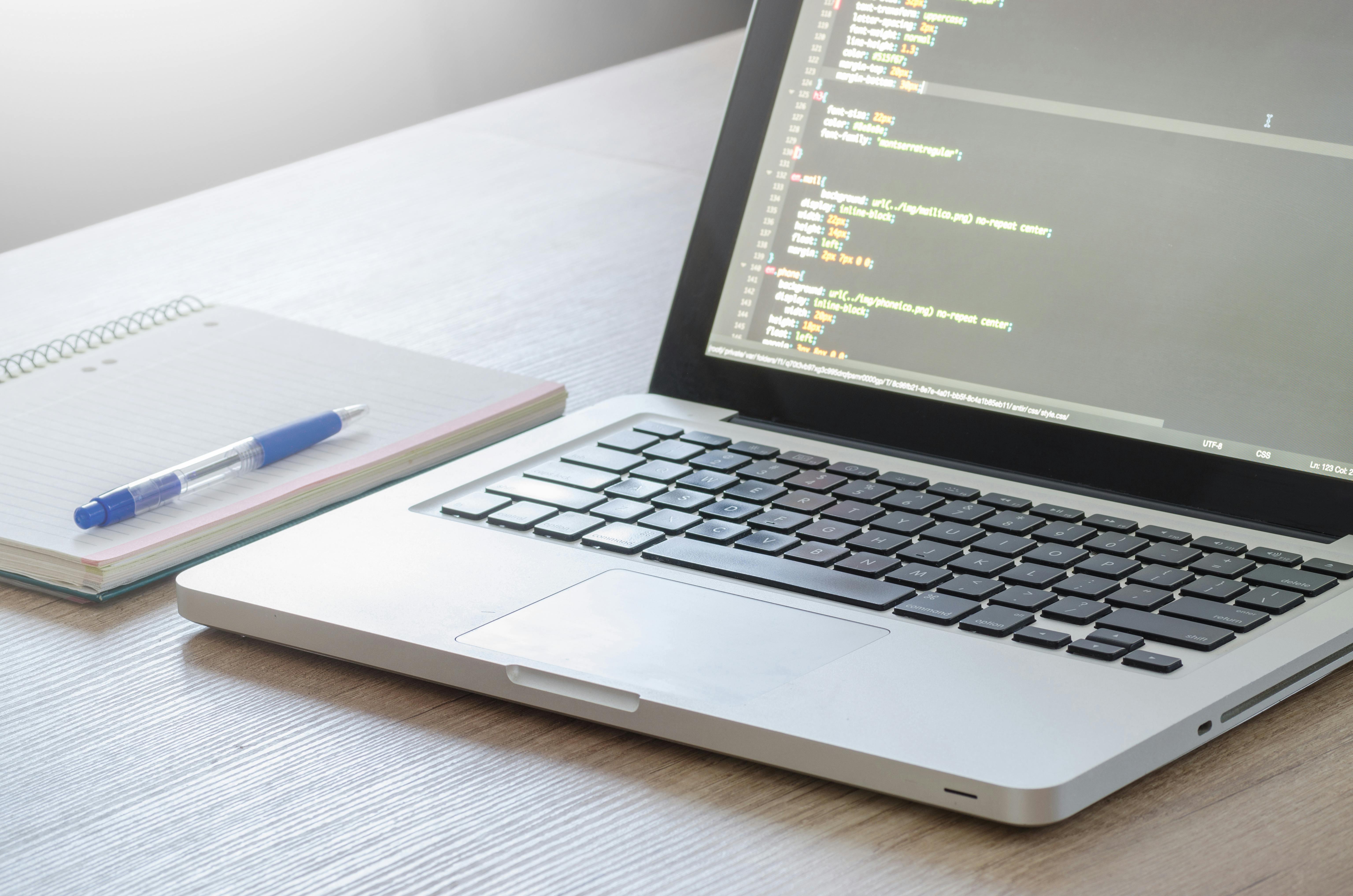 Open laptop displaying code on screen next to a spiral notebook and pen on a wooden desk in a bright, calm workspace