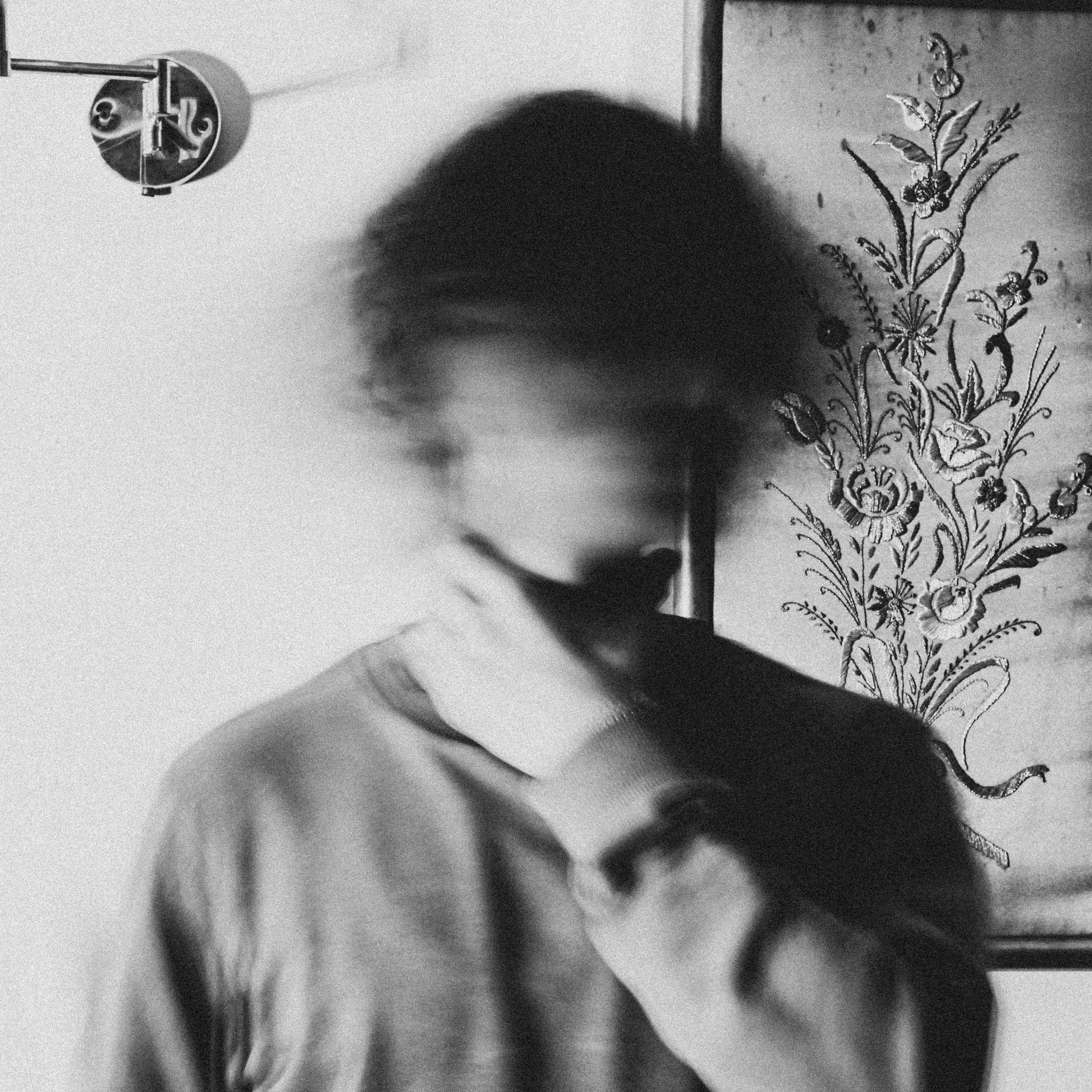 Person standing indoors with head and face blurred in motion, wearing a long-sleeve shirt and touching their neck, next to a wall with a decorative floral artwork and a modern wall lamp, overall mood is introspective and mysterious